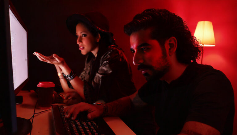 Dois editores de vídeo, um homem e uma mulher, olham perplexos para o monitor. Ambiente com luz vermelha dramática.